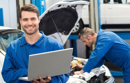 A mechanic holding a laptop smiling at a camera, a second mechanic is working on a car in the background